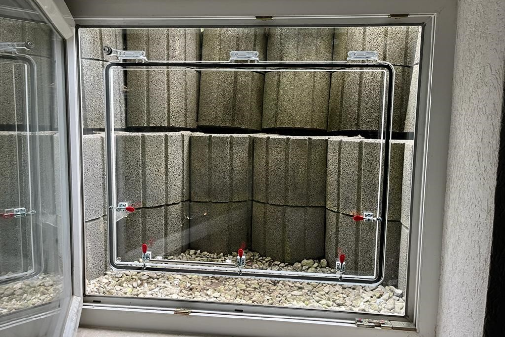 AquaLOCK® Hochwasserschutzfenster mit Schutzrahmen und Blick in einen Lichtschacht aus Steinen